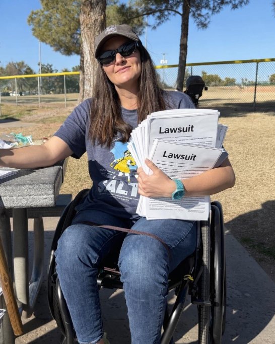 Gina Schuh outdoors at a picnic table in her wheelchair, a thick stack of lawsuit filings under one arm and a pitchfork in her other hand.